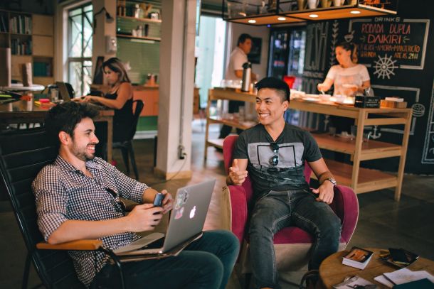 Imagem de dois homens sentados em uma mesa de coworking, interagindo entre si com um sorriso enquanto usam seus laptops. Um ambiente casual e moderno de trabalho colaborativo ao fundo sugere um equilíbrio entre qualidade de vida e produtividade