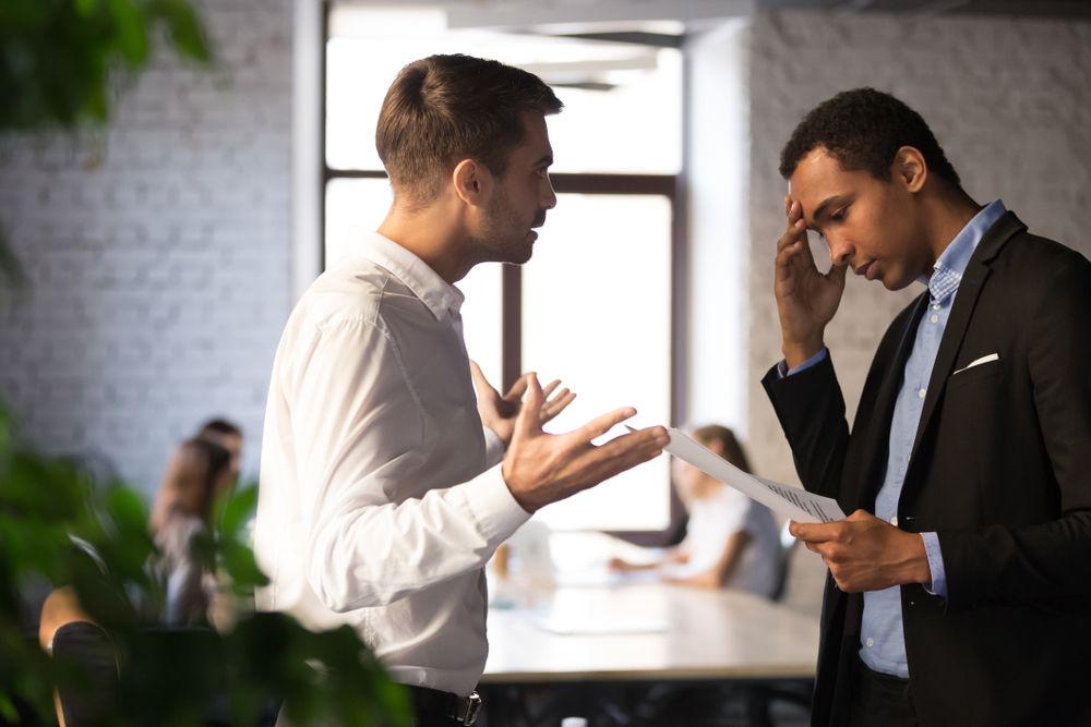 Na imagem, observamos um momento de interação entre dois líderes no ambiente de trabalho. O homem à esquerda, vestido com uma camisa branca, está gesticulando e parece estar explicando um ponto importante, possivelmente oferecendo conselhos ou soluções. O homem à direita, usando um blazer escuro e camisa azul com colarinho aberto, segura um documento e tem uma expressão de preocupação ou reflexão, com a mão na testa, indicando que ele está processando a informação. A cena sugere um diálogo onde a liderança colaborativa está em prática, destacando a importância do suporte mútuo e da comunicação aberta entre líderes para o sucesso organizacional e pessoal.