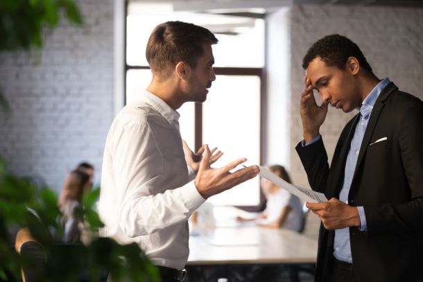 Na imagem, observamos um momento de interação entre dois líderes no ambiente de trabalho. O homem à esquerda, vestido com uma camisa branca, está gesticulando e parece estar explicando um ponto importante, possivelmente oferecendo conselhos ou soluções. O homem à direita, usando um blazer escuro e camisa azul com colarinho aberto, segura um documento e tem uma expressão de preocupação ou reflexão, com a mão na testa, indicando que ele está processando a informação. A cena sugere um diálogo onde a liderança colaborativa está em prática, destacando a importância do suporte mútuo e da comunicação aberta entre líderes para o sucesso organizacional e pessoal.