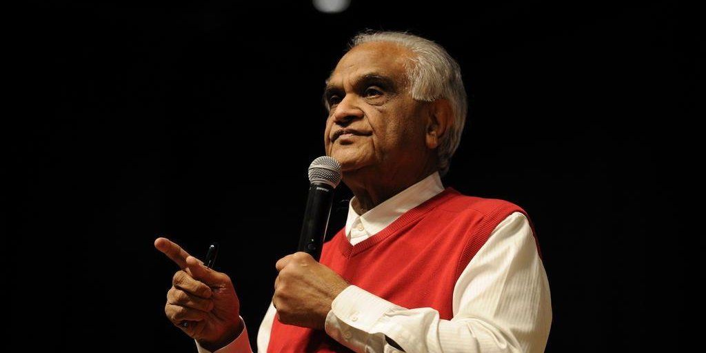 A imagem mostra Ram Charan, um homem de meia idade de origem indiana, com vestes formais tradicionais e em uma palestra, segurando o microfone em uma mão e apontando para a plateia com a outra.